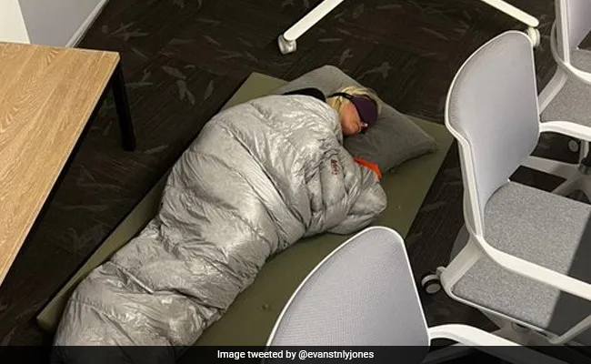 A woman sleeping on the ground of her office in a sleeping bag with an eye mask.